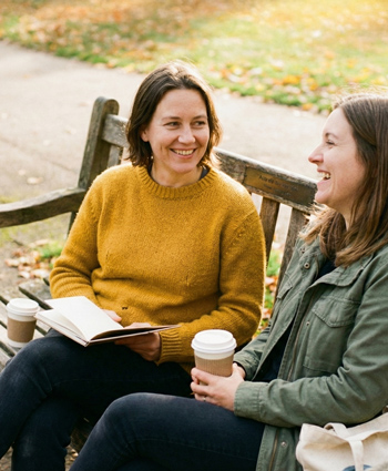 Two women sit on a bench in a park, holding to-go coffee cup, talking and laughing.